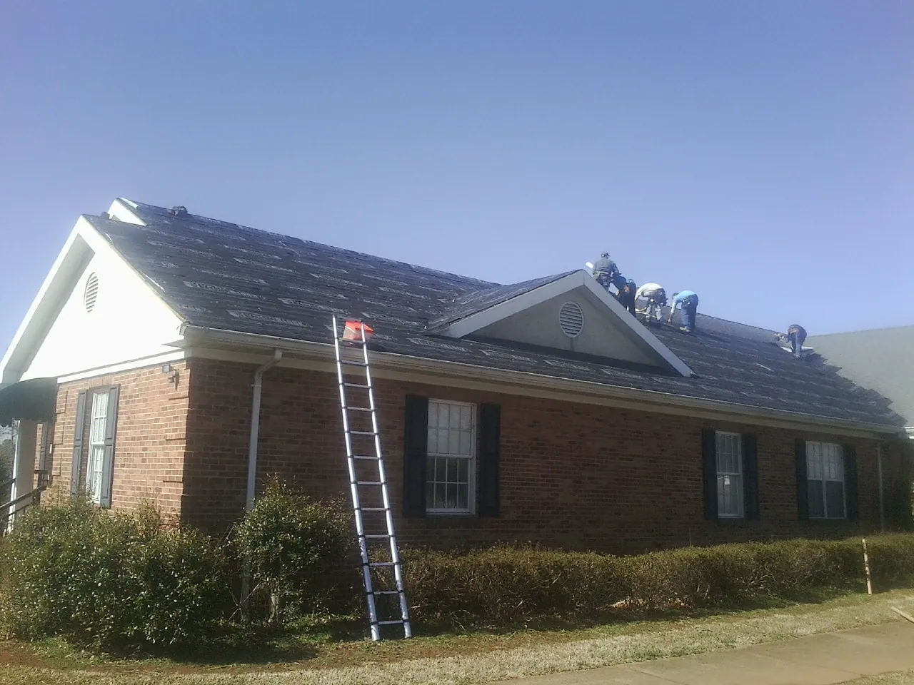 Crew tearing shingles off a brick ranch roof with a ladder set — Metal Roof Repair work by our crew in New Site