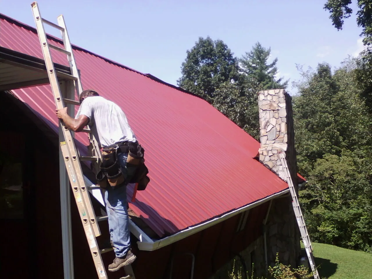 Roofer on a ladder beside a red metal roof and stone chimney — Soffit & Fascia Repair by Roofing Brothers in New Site